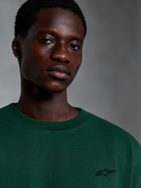 Alpinestars Essentials Ageless SS Tee, maglietta a maniche corte verde pino, primo piano di un modello che indossa una t-shirt in cotone organico verde con piccolo logo ricamato, design streetwear casual