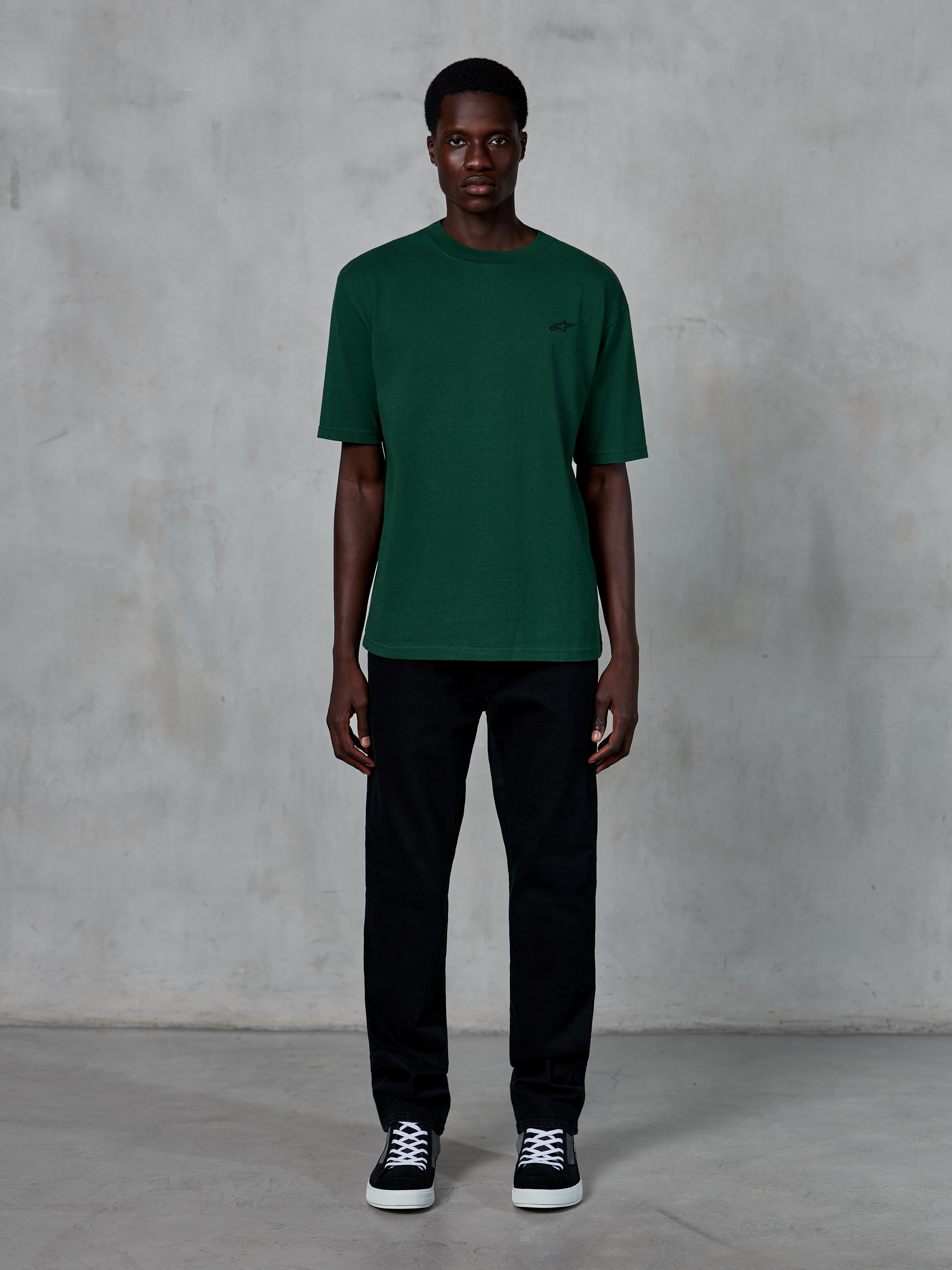 Alpinestars Essentials Ageless SS Tee, maglietta a maniche corte, verde pino, con un discreto logo nero sul petto, indossata da un modello con pantaloni neri e sneakers, streetwear casual lifestyle con vestibilità rilassata