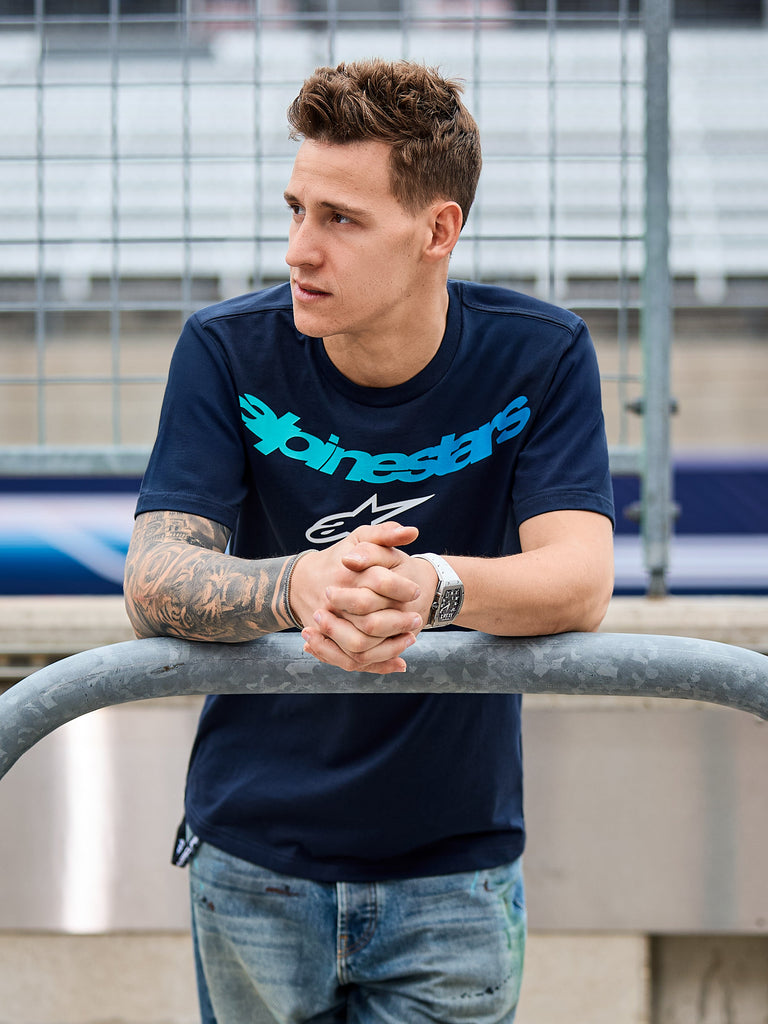 Alpinestars Collared CSF Tee, t-shirt a maniche corte, blu navy, scritta Alpinestars color ottanio e logo a stella bianco, indossata da un uomo appoggiato a una ringhiera di metallo in un circuito, streetwear casual motorsport
