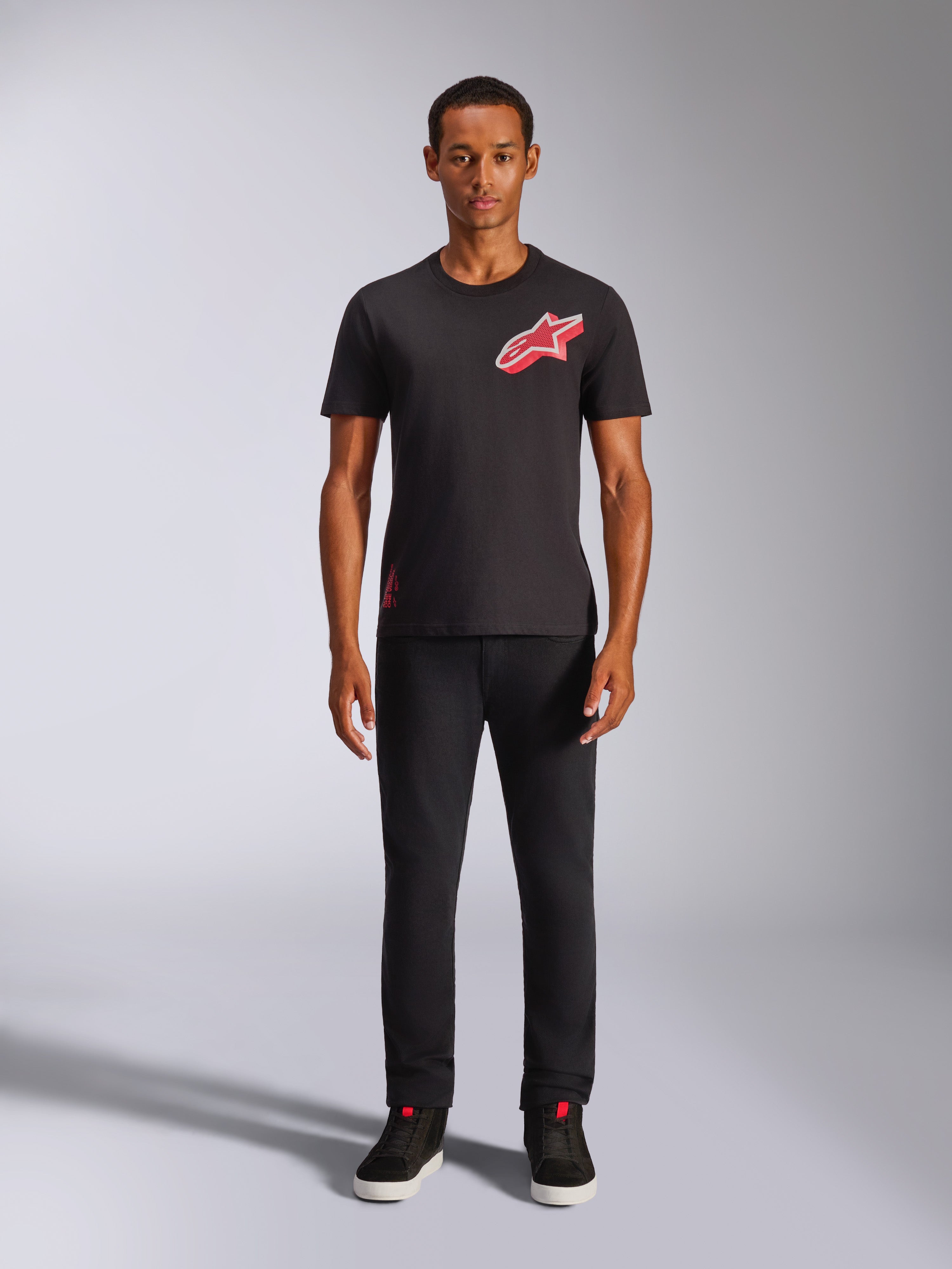 Modello che indossa la t-shirt Alpinestars Shaded CSF a maniche corte nera, con logo Astars rosso sul petto caratterizzato da stampa a gradiente in lamina inclinata, abbinata a pantaloni neri e sneakers, streetwear casual motorsport.