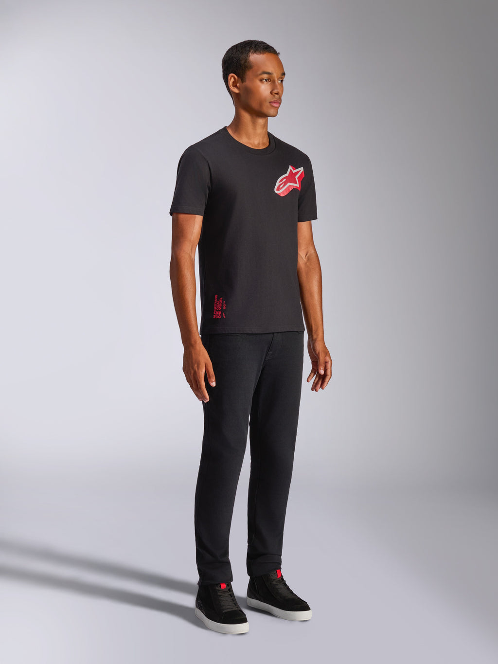 Alpinestars Shaded CSF Tee, maglietta a maniche corte, nero, logo Astars con stampa a gradiente in lamina inclinata rosso e bianco, streetwear casual motorsport, inquadratura a figura intera di un modello che indossa pantaloni neri e sneaker
