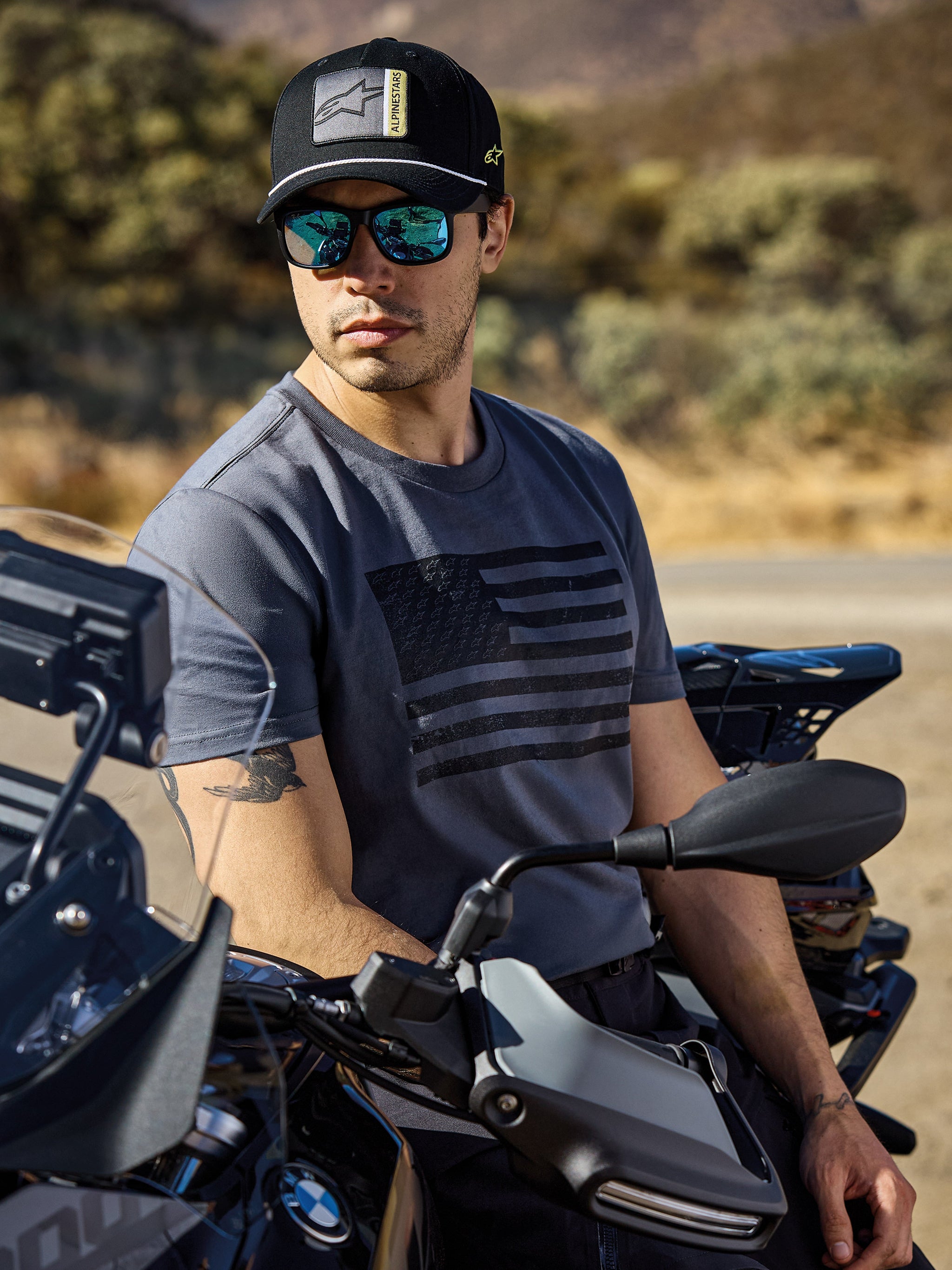 Alpinestars Waving CSF Tee, maglietta a maniche corte, Charcoal, con grafica a bandiera nera sul petto, indossata da un uomo seduto su una moto in un ambiente esterno soleggiato, stile di vita casual motorsport