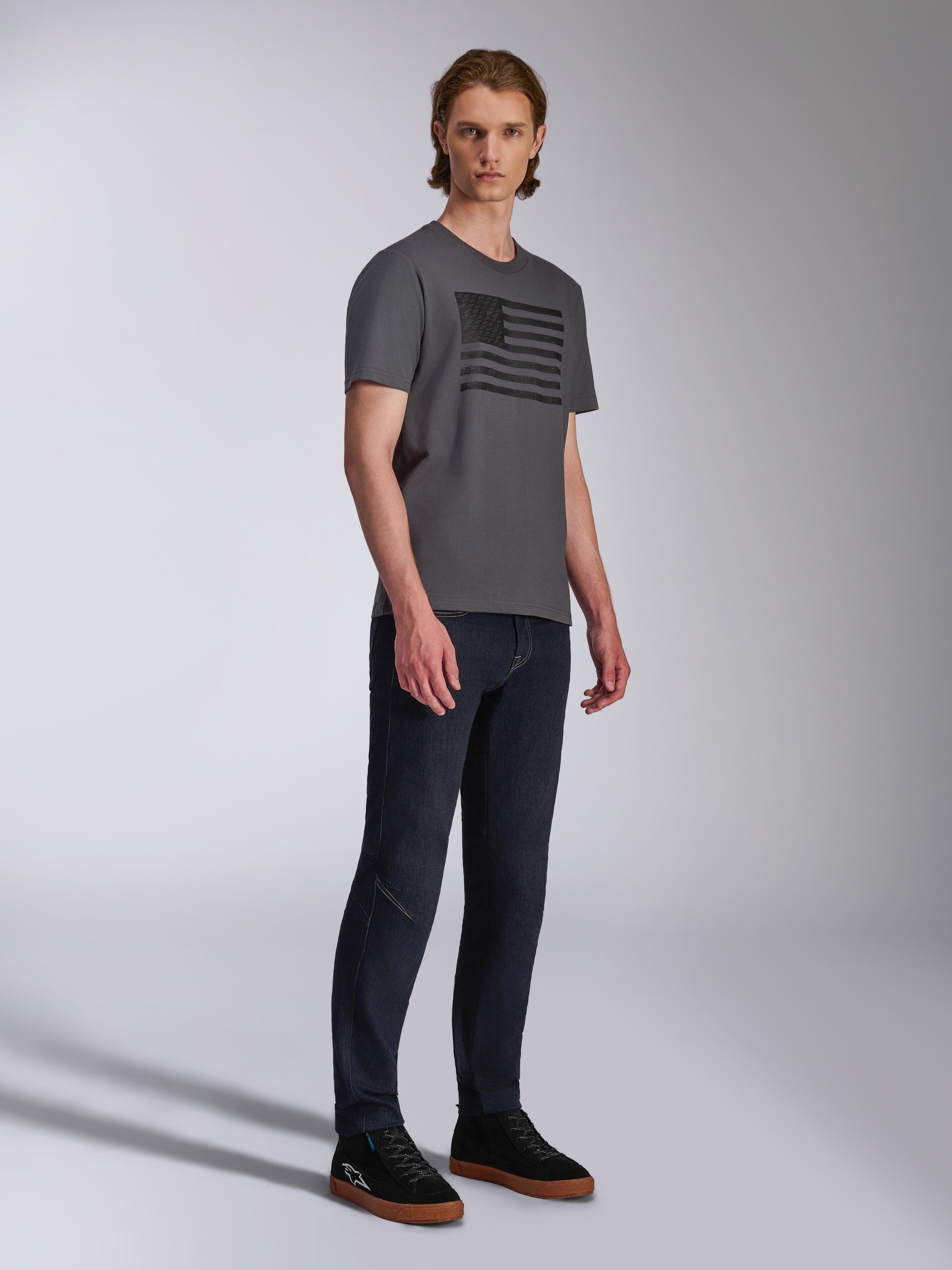 Alpinestars Waving CSF Tee, maglietta a maniche corte, antracite, grafica con bandiera ondulata nera, indossata da un modello con jeans scuri e sneakers, abbigliamento streetwear casual motorsport