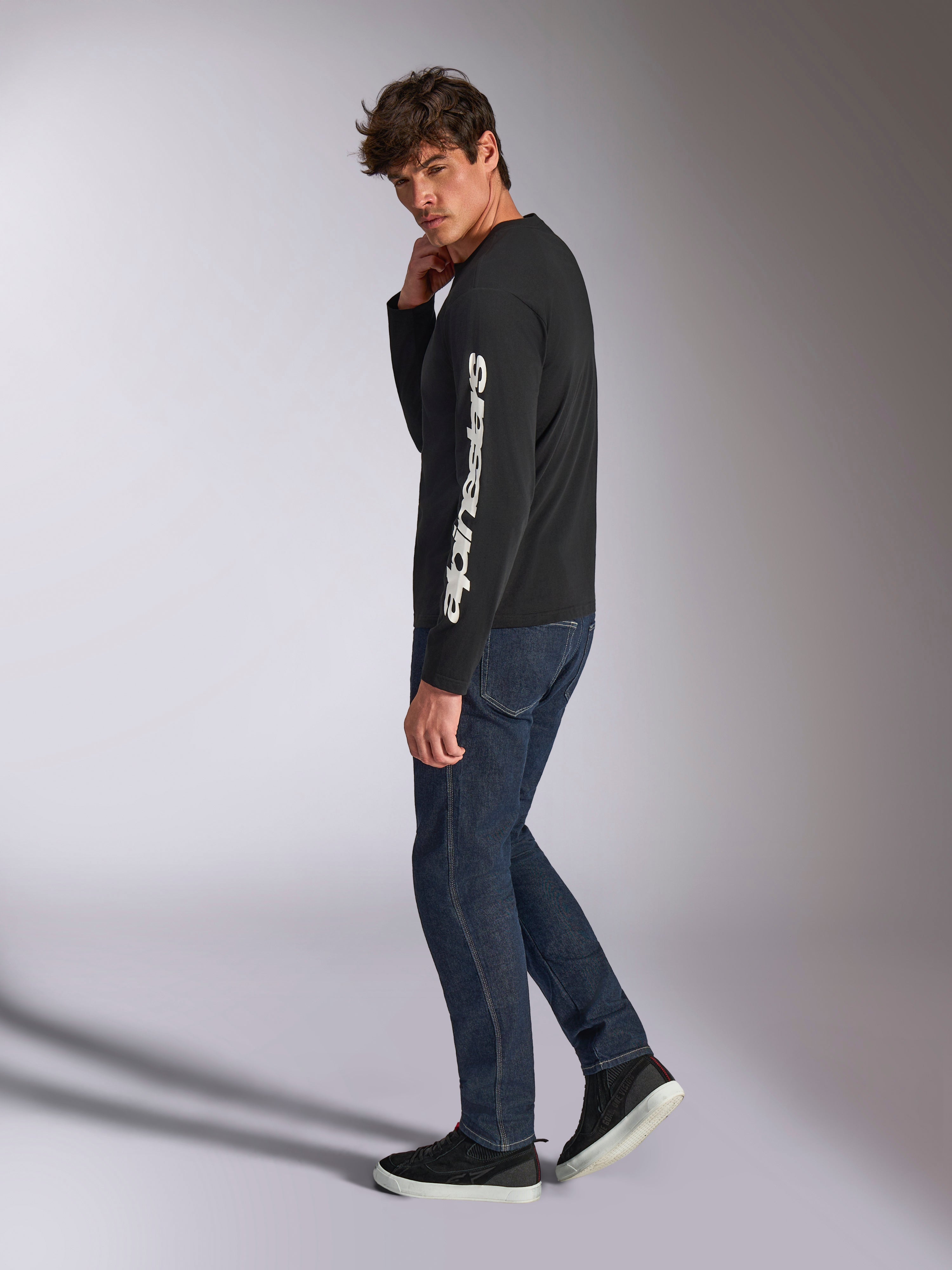 Alpinestars Trackside CSF Long Sleeve Tee, maglia a maniche lunghe, nera con scritta Alpinestars verticale bianca sulla manica, indossata da un modello con denim scuro e sneakers, abbigliamento sportivo casual ispirato al motorsport