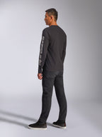 Alpinestars Trackside CSF Long Sleeve Tee, maglia a maniche lunghe nera, con grande logo Alpinestars grigio sulla manica sinistra, indossata da un modello con pantaloni neri e sneakers casual, streetwear casual ispirato al motorsport