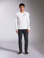 Alpinestars Trackside CSF Tee a maniche lunghe, bianca con branding nero, piccolo logo sul petto e scritta verticale sulla manica, indossata da un modello con pantaloni scuri e sneakers nere, streetwear casual ispirato al motorsport