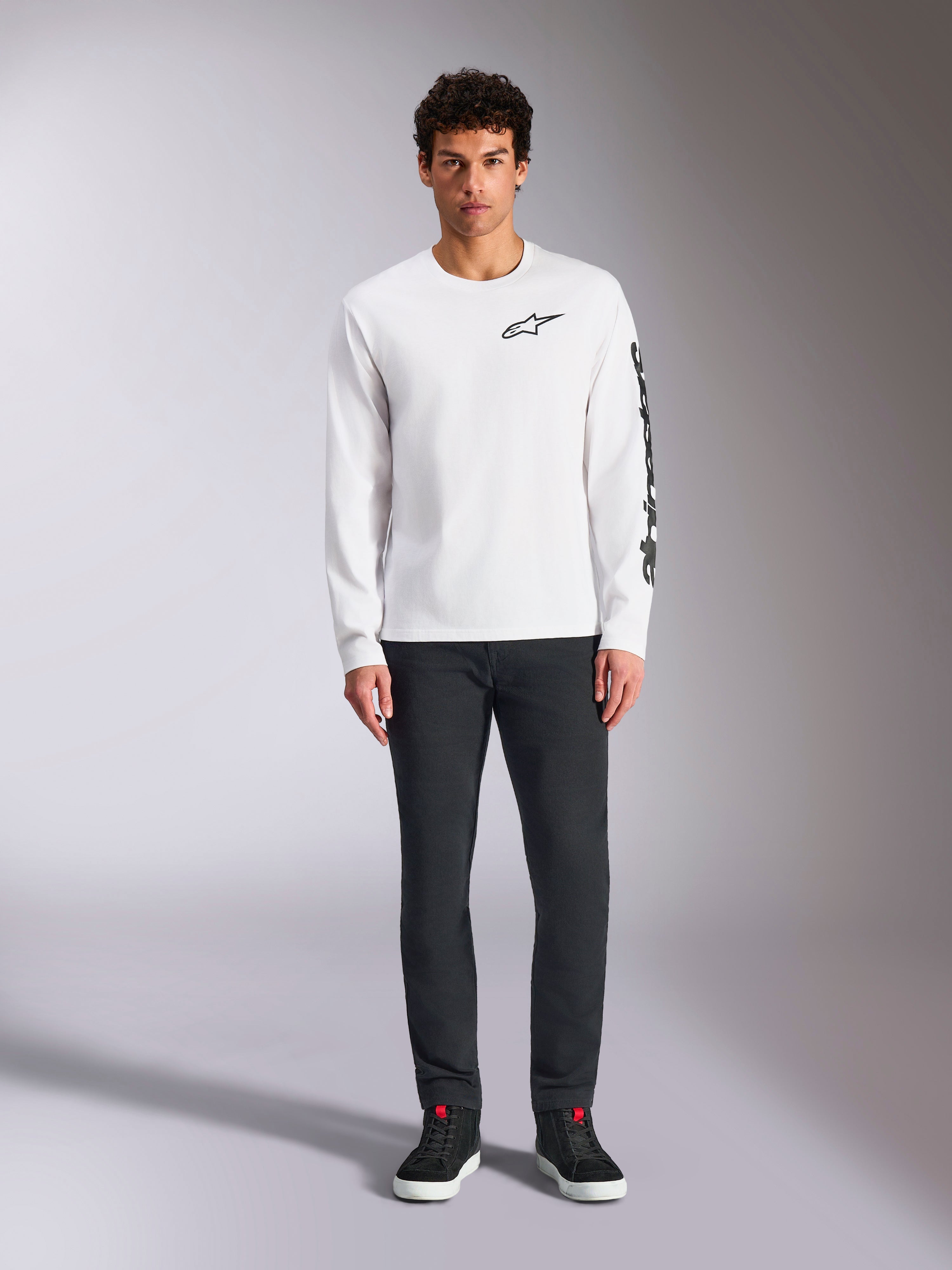 Alpinestars Trackside CSF Tee a maniche lunghe, bianca con branding nero, piccolo logo sul petto e scritta verticale sulla manica, indossata da un modello con pantaloni scuri e sneakers nere, streetwear casual ispirato al motorsport
