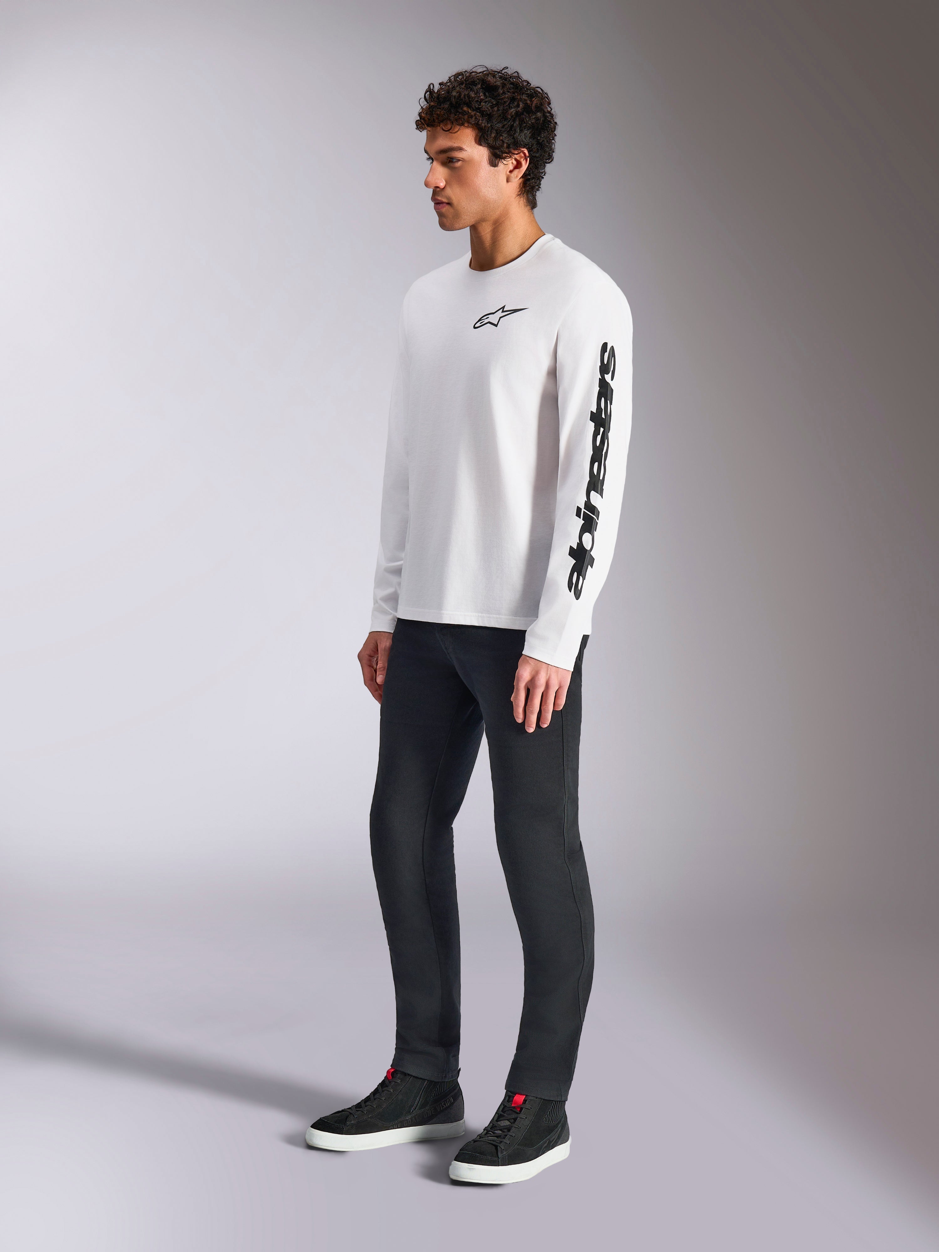 Alpinestars Trackside CSF Tee, maglietta a maniche lunghe, bianca, logo a stella nero sul petto e scritta sulla manica sinistra, indossata da un modello con pantaloni scuri e sneakers, abbigliamento sportivo casual lifestyle