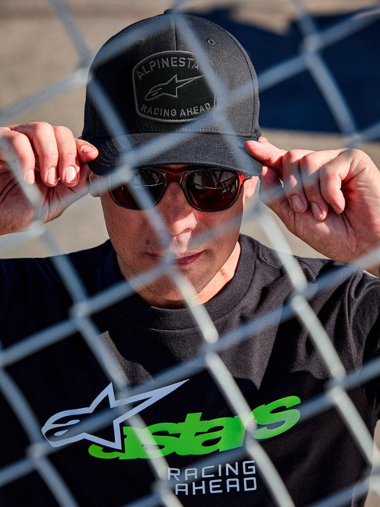 Cappello Alpinestars Promptus Snapback, berretto lifestyle nero, visiera piatta con corona strutturata, patch frontale ricamata curva con cordina intrecciata, indossato da un uomo con occhiali da sole e t-shirt nera con logo Astars verde, scatto attraverso una recinzione a maglie metalliche