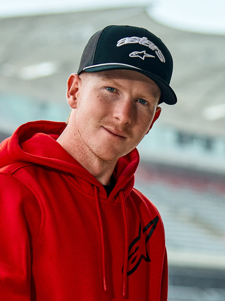 Uomo con felpa rossa Alpinestars e cappello Agree Trucker nero, loghi Astars ricamati in bianco, dettaglio con corda intrecciata sulla visiera e retro in rete traspirante, stile lifestyle casual