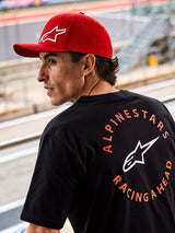 Alpinestars Corp Shift Multi Hat, cappello rosso con visiera curva e logo Astars 3D bianco, indossato con una t-shirt nera Alpinestars con grafica 'Racing Ahead', ambientazione motorsport lifestyle in un circuito di gara