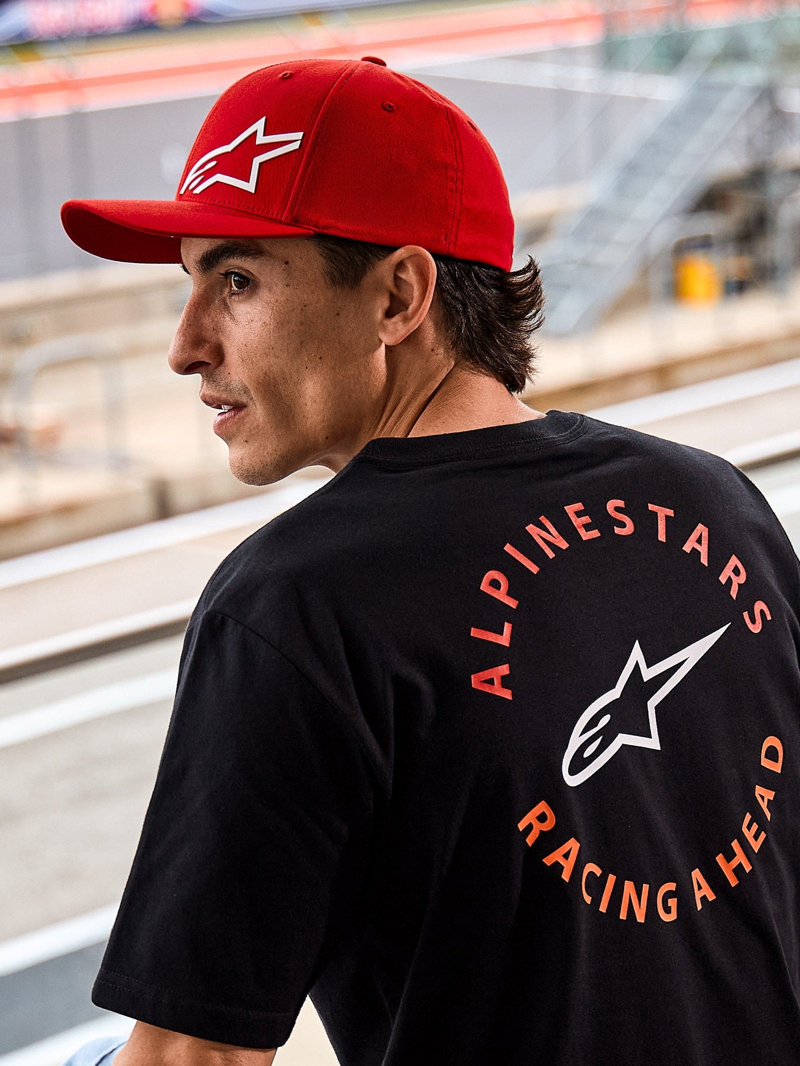 Alpinestars Corp Shift Multi Hat, cappello rosso con visiera curva e logo Astars 3D bianco, indossato con una t-shirt nera Alpinestars con grafica 'Racing Ahead', ambientazione motorsport lifestyle in un circuito di gara