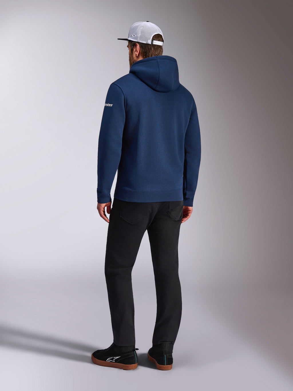 Alpinestars Frontal Hoodie, vista posteriore di una felpa in pile con cappuccio blu navy, logo con scritta bianco sulla manica, abbinata a pantaloni neri e un cappellino trucker in mesh bianco, streetwear casual ispirato al motorsport
