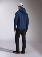 Alpinestars Frontal Hoodie, vista posteriore di una felpa in pile con cappuccio blu navy, logo con scritta bianco sulla manica, abbinata a pantaloni neri e un cappellino trucker in mesh bianco, streetwear casual ispirato al motorsport
