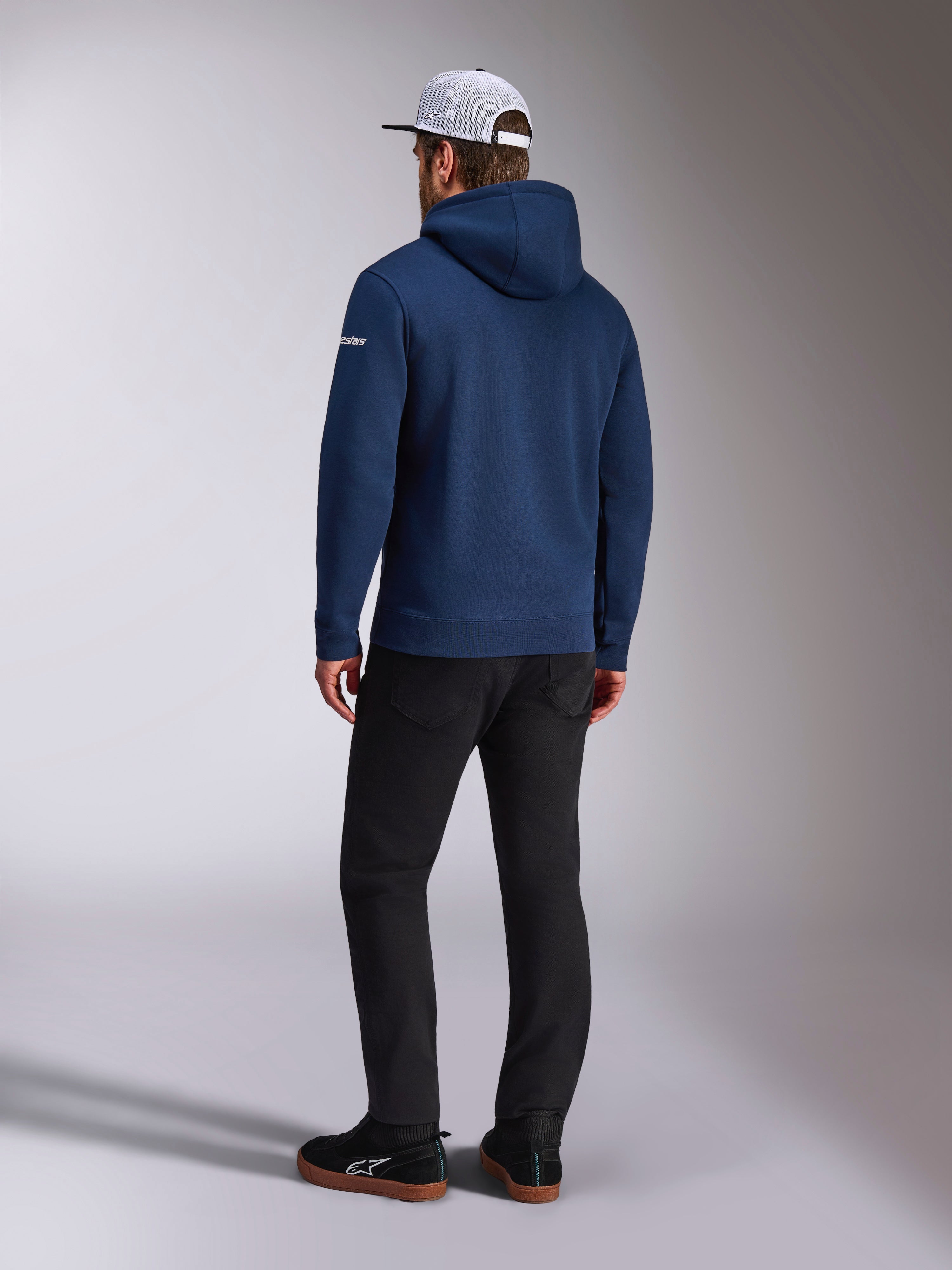 Alpinestars Frontal Hoodie, vista posteriore di una felpa in pile con cappuccio blu navy, logo con scritta bianco sulla manica, abbinata a pantaloni neri e un cappellino trucker in mesh bianco, streetwear casual ispirato al motorsport
