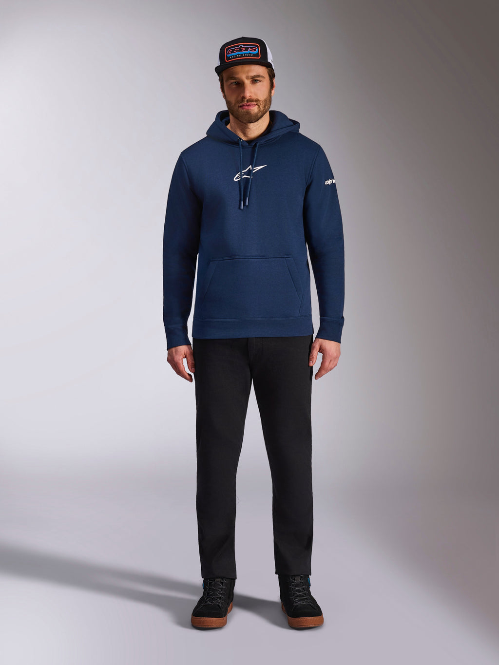 Modello indossa la felpa con cappuccio Alpinestars Frontal in colore Navy, felpa in pile con logo Astars bianco sul petto e logo con scritta sulla manica, con tasca a marsupio e cappuccio con cordino, abbinata a pantaloni neri e cappellino con logo, streetwear casual motorsport