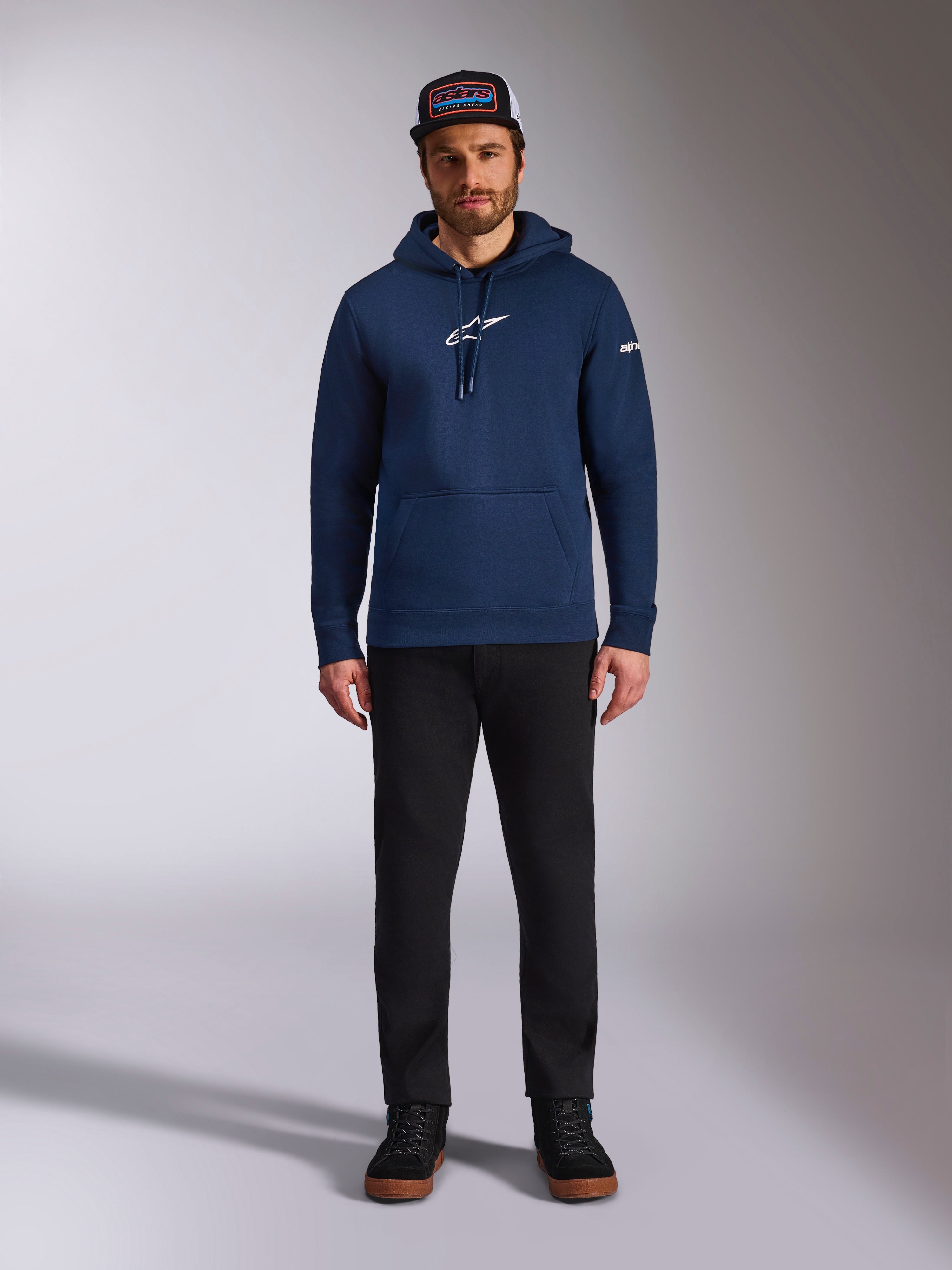 Modello indossa la felpa con cappuccio Alpinestars Frontal in colore Navy, felpa in pile con logo Astars bianco sul petto e logo con scritta sulla manica, con tasca a marsupio e cappuccio con cordino, abbinata a pantaloni neri e cappellino con logo, streetwear casual motorsport