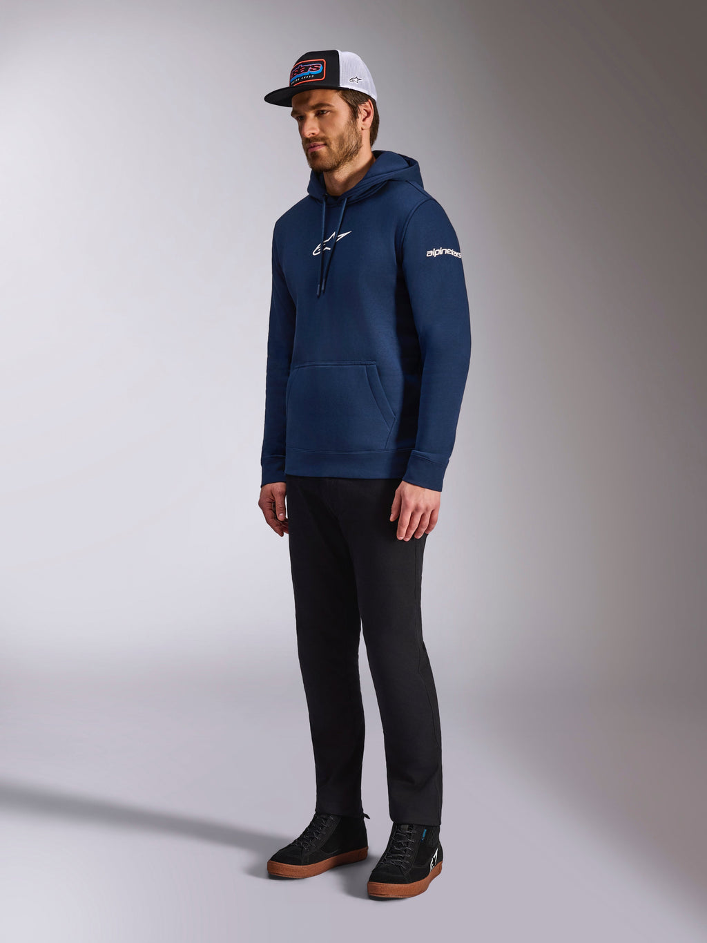Felpa con cappuccio Alpinestars Frontal in blu navy, felpa casual in pile con logo Astars bianco sul petto e scritta sulla manica, abbinata a un cappellino trucker e pantaloni neri, abbigliamento lifestyle moderno d'ispirazione motorsport.