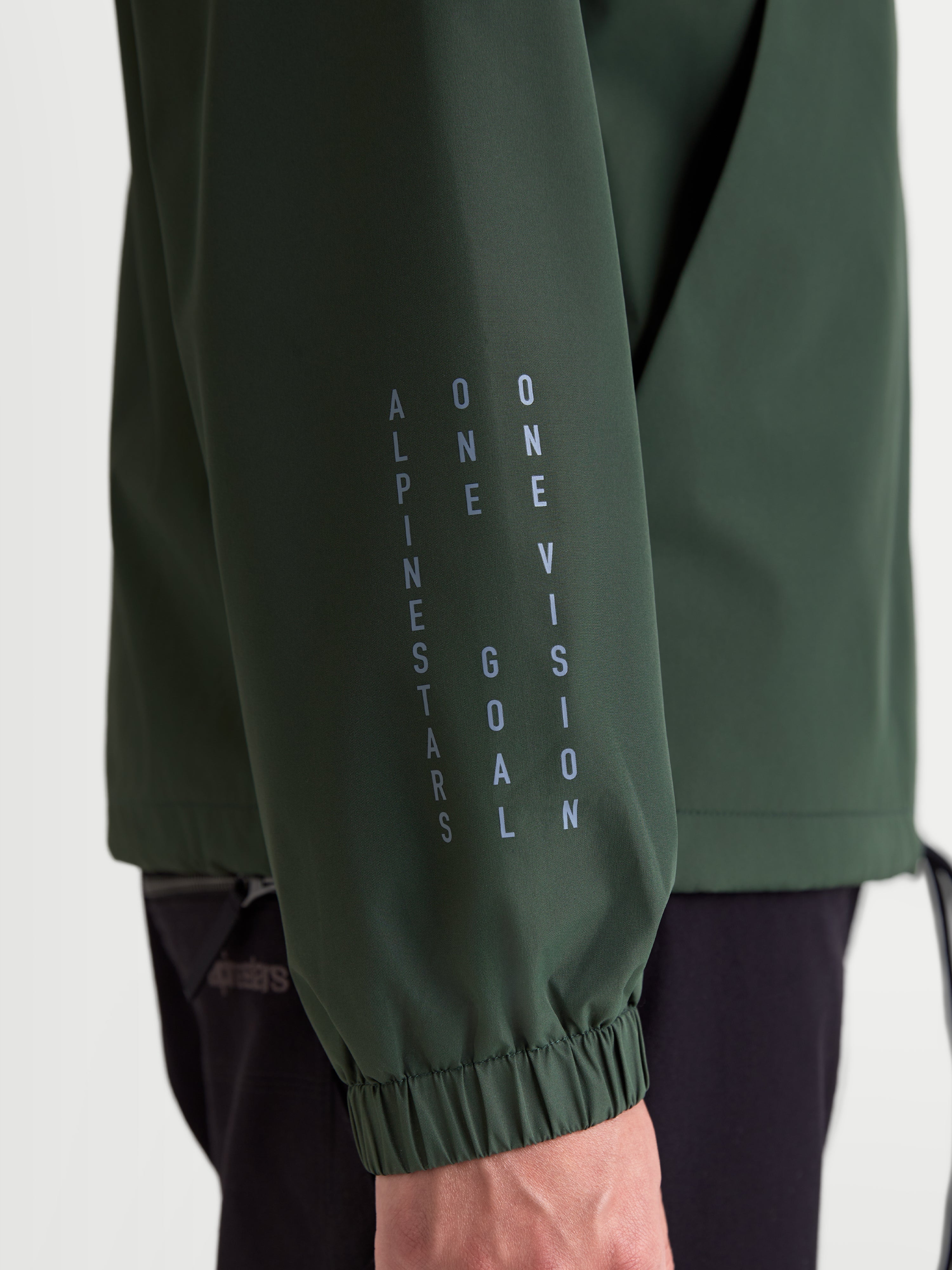 Alpinestars Giacca Frangivento Speeder Resolve, giacca frangivento sportswear leggera, verde pino, dettaglio ravvicinato della manica con marchio Alpinestars in verticale, motto One Goal One Vision e polsino elastico.