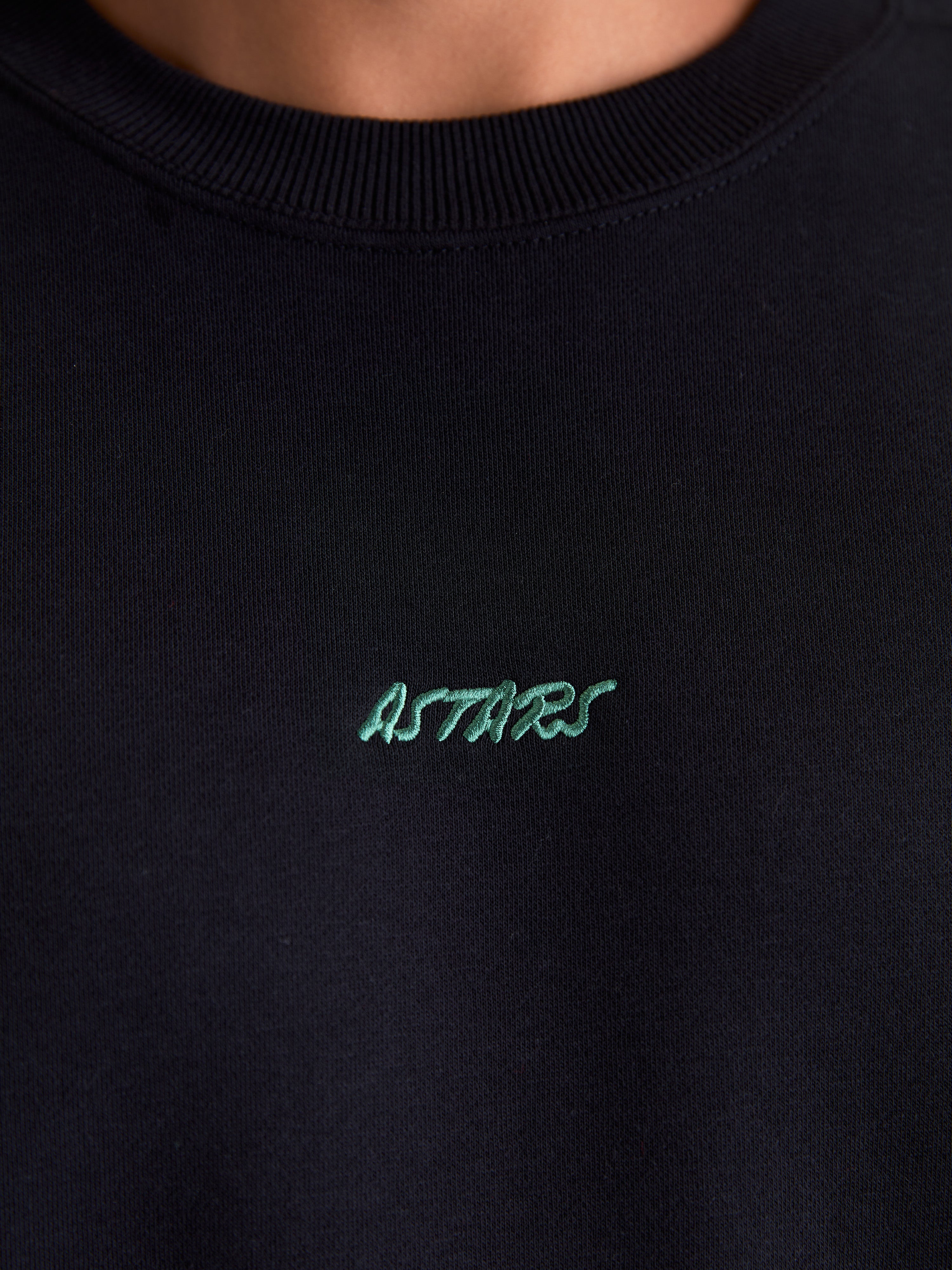 Alpinestars Guise Oversized Crew, felpa girocollo sportswear in pile, nera, primo piano del colletto a costine e logo Astars ricamato in verde sul petto, realizzata in misto cotone e poliestere riciclato