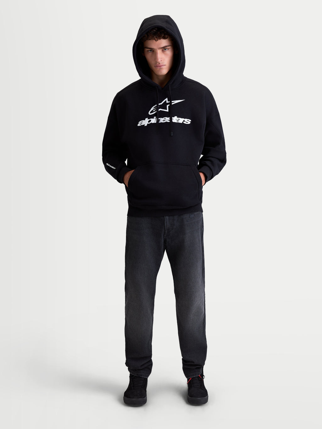 Alpinestars Felpa con cappuccio Prelude Oversized, felpa sportswear da uomo, nera e bianca, vestibilità oversize con tasca a marsupio e loghi con stampa a rilievo, in cotone e poliestere riciclato, indossata da un modello.