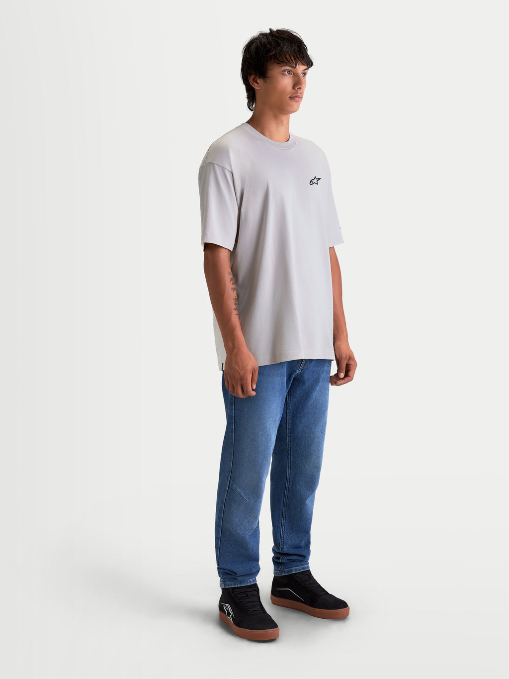 Alpinestars maglietta Mandate Oversized CSF, maglietta sportswear a maniche corte unisex, grigio cemento, vestibilità oversized rilassata con piccolo logo Astars nero sul petto. Capo in misto cotone e poliestere per il massimo comfort casual, indossata da un modello con jeans blu e sneakers nere.