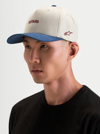 Alpinestars Cappello County Canvas, cappello sportswear in sabbia e blu, indossato da un modello, con visiera curva e logo Astars ricamato