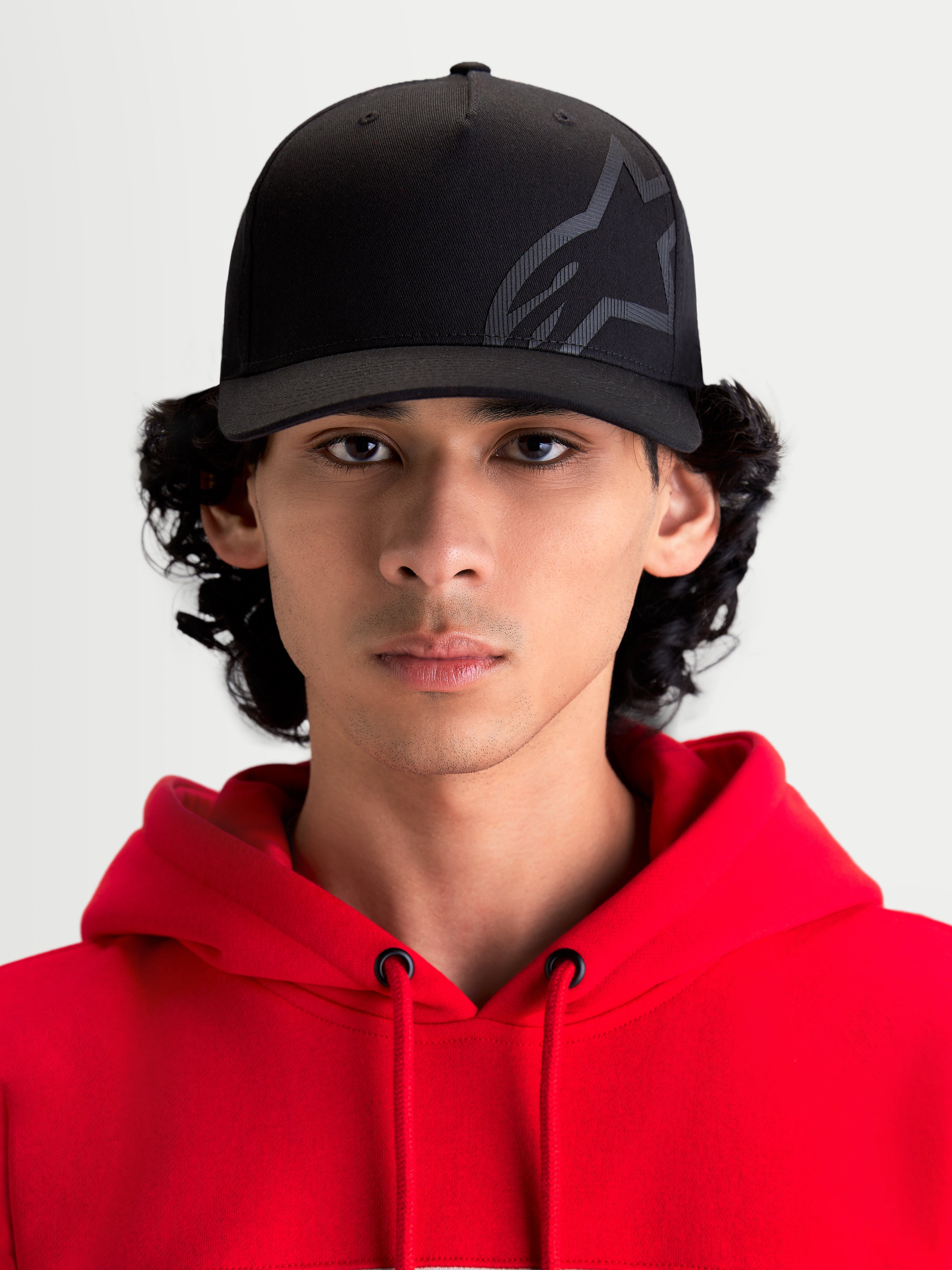 Alpinestars Cappello Maze, cappello sportswear ispirato al motorsport, nero, indossato da un modello con felpa con cappuccio rossa, corona strutturata, visiera curva, logo Astars Corp Shift testurizzato in tessuto twill resistente, stile casual off-bike
