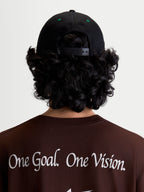 Alpinestars Cappello Retro Snapback, cappello snapback sportswear, nero e verde, vista posteriore su modello con chiusura regolabile e occhielli verdi, indossato con maglietta marrone con il manifesto Alpinestars One Goal. One Vision.