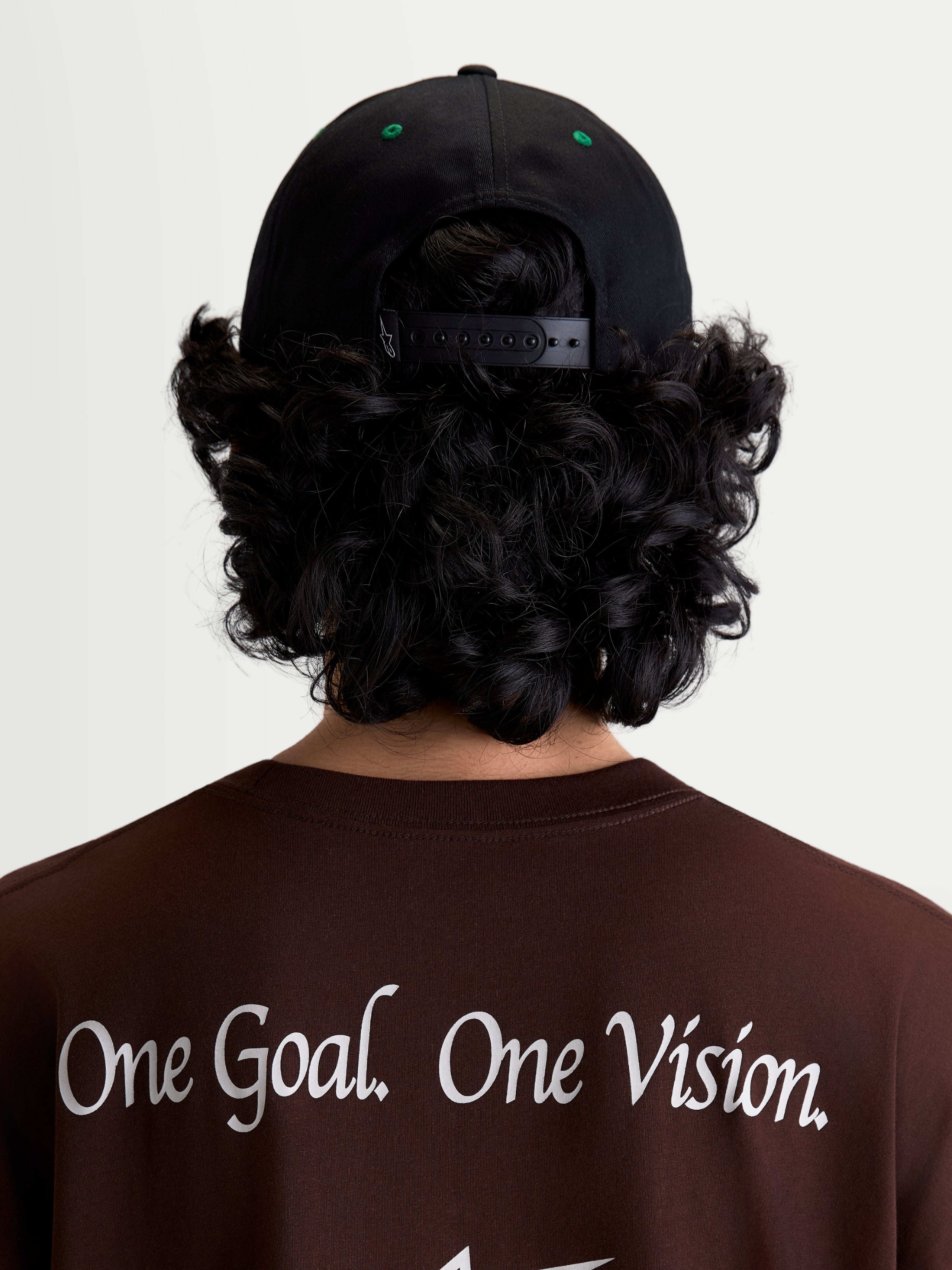 Alpinestars Cappello Retro Snapback, cappello snapback sportswear, nero e verde, vista posteriore su modello con chiusura regolabile e occhielli verdi, indossato con maglietta marrone con il manifesto Alpinestars One Goal. One Vision.