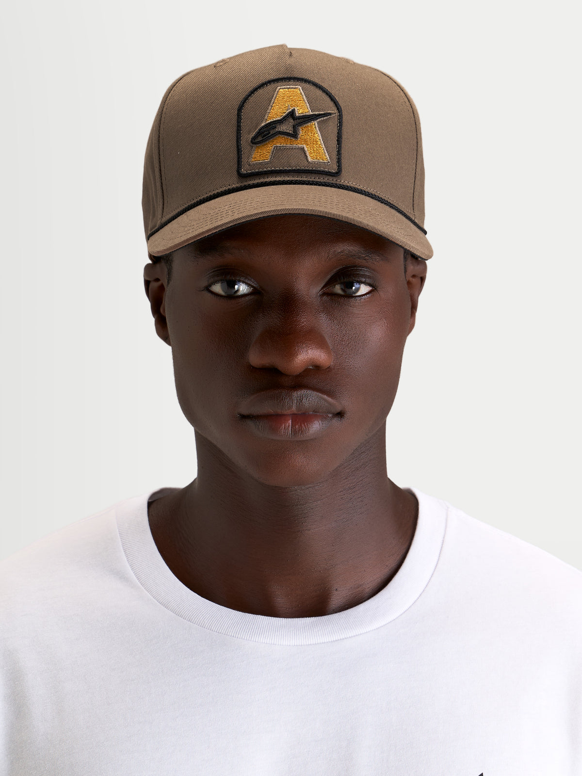 Alpinestars Cappello Acme Snapback, cappello sportswear marrone indossato da un modello, con corona strutturata, visiera curva e patch frontale ricamata con logo Astars e rifinitura in corda