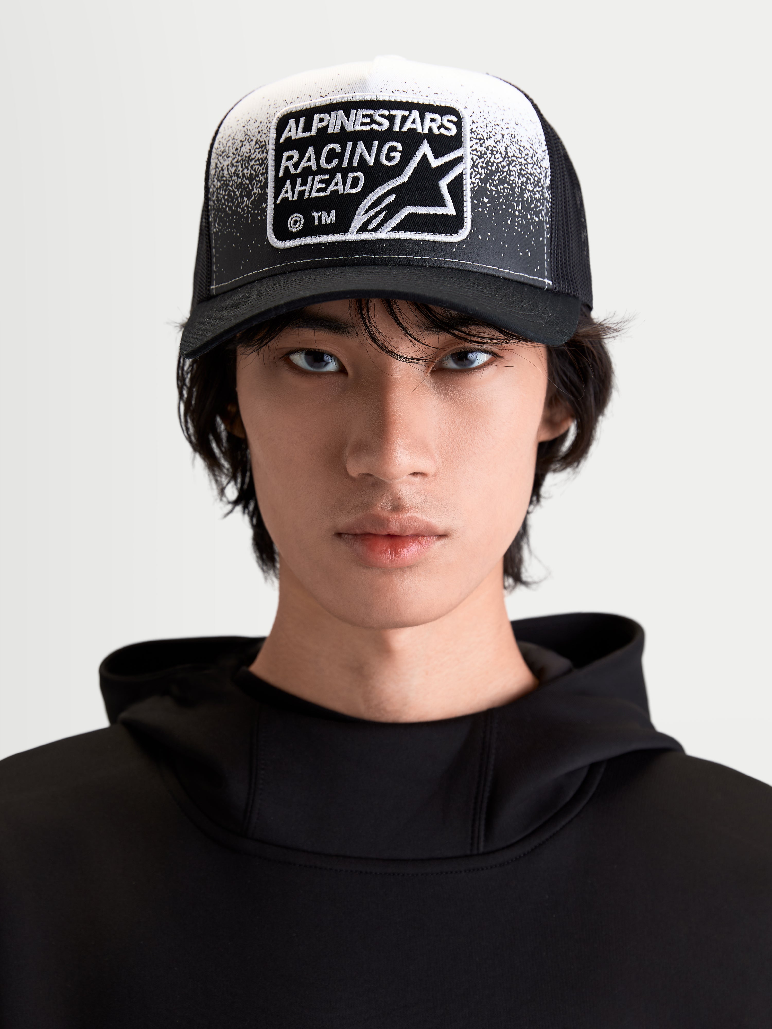 Alpinestars Cappello Gravel, cappello trucker sportswear lifestyle unisex, nero e bianco, vista frontale indossata con visiera curva, retro in rete traspirante e patch Racing Ahead con texture effetto ghiaia