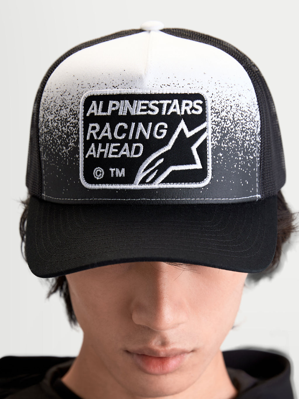 Alpinestars cappello Gravel, cappello trucker sportswear unisex, nero e bianco, con patch tessuta Alpinestars Racing Ahead e dettaglio effetto ghiaia, corona strutturata e visiera curva, vista frontale su modello