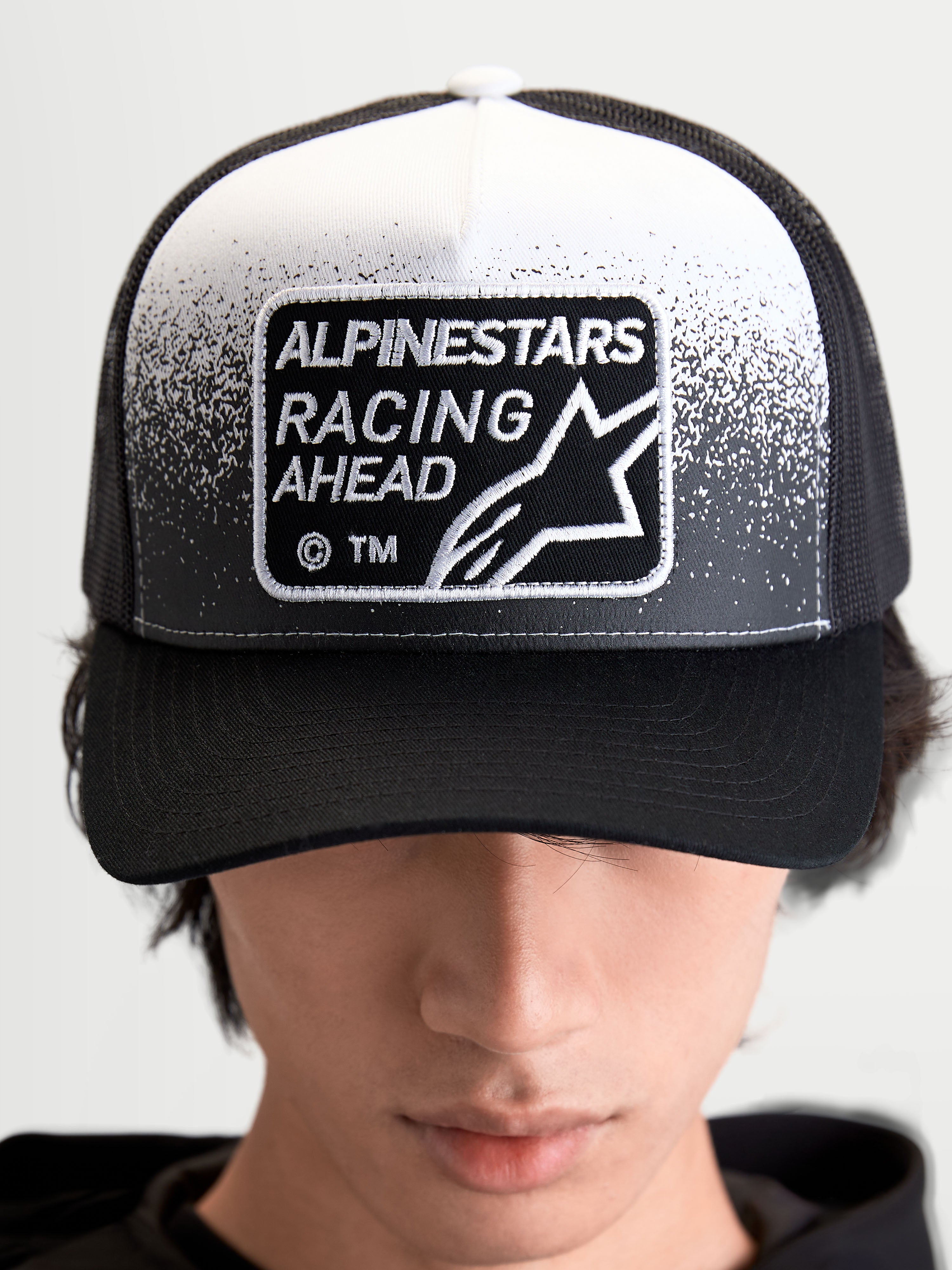 Alpinestars cappello Gravel, cappello trucker sportswear unisex, nero e bianco, con patch tessuta Alpinestars Racing Ahead e dettaglio effetto ghiaia, corona strutturata e visiera curva, vista frontale su modello