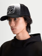 Alpinestars Cappello Gravel, cappello trucker casual indossato da un modello, nero e bianco, con corona strutturata, visiera curva e retro in rete traspirante. Caratterizzato da patch frontale con grafica effetto ghiaia e marchio Alpinestars Racing Ahead.