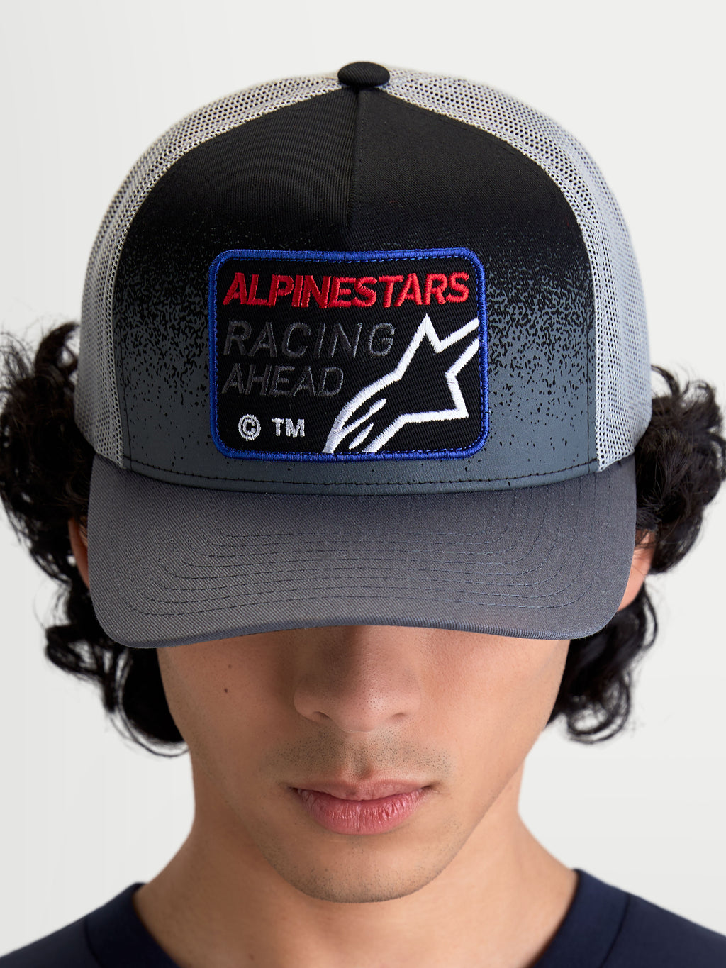 Alpinestars Cappello Gravel Trucker, cappello trucker sportswear, grigio scuro e nero, visiera curva, corona strutturata, patch frontale tessuta con logo Astars e scritta Racing Ahead, retro in rete traspirante, indossato dal modello vista frontale