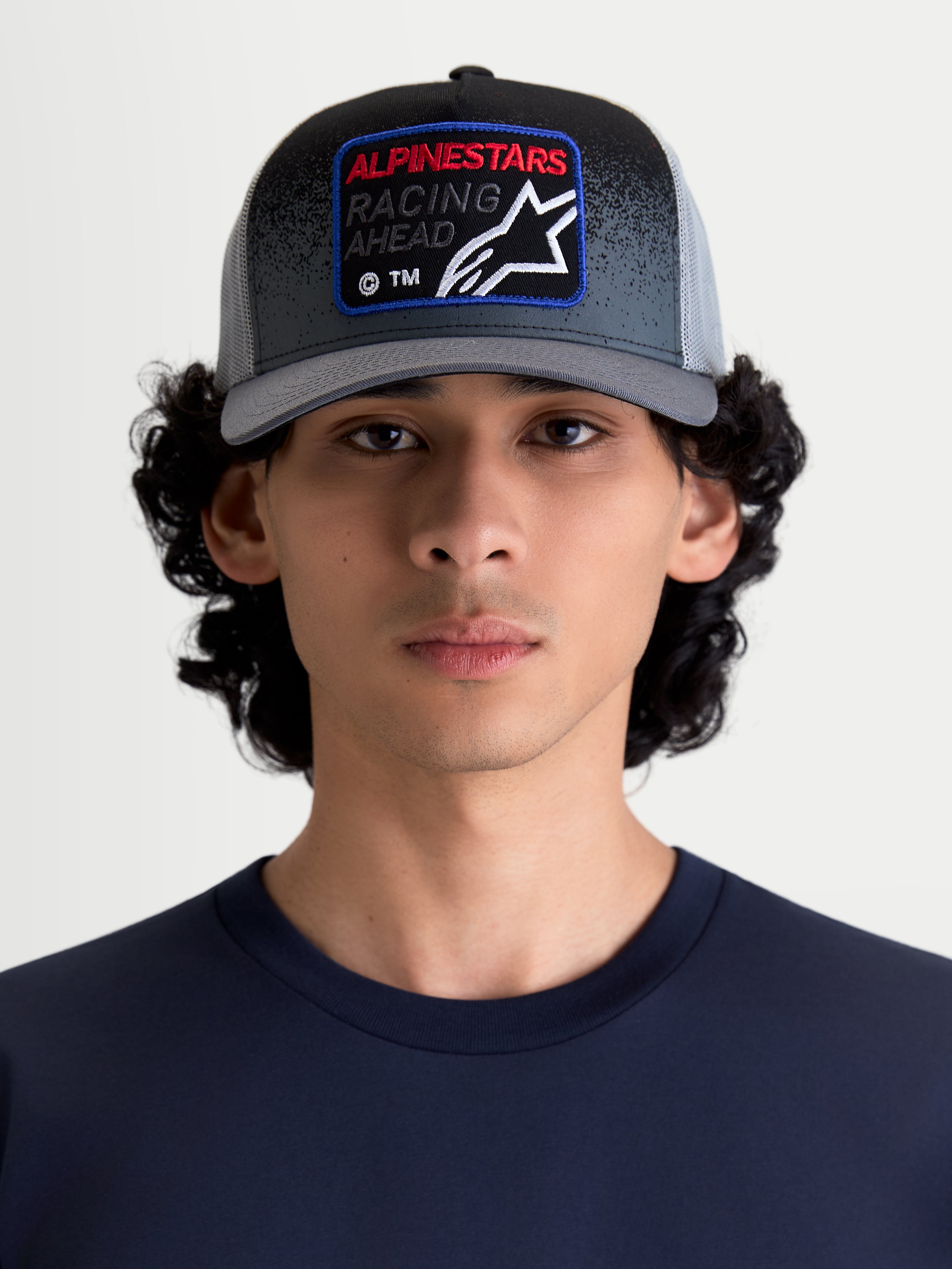 Alpinestars Cappello Gravel Trucker, cappello sportswear casual con retro in rete per uomo e donna, grigio scuro e nero, indossato da un modello, con corona strutturata, visiera curva e patch frontale tessuta Alpinestars Racing Ahead con dettagli effetto ghiaia.