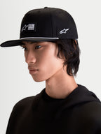 Alpinestars Cappello Label Snapback, cappello sportivo con visiera piatta e corona strutturata, nero con dettagli bianchi, etichetta tessuta frontale e cordino a contrasto, indossato da un modello per uno stile lifestyle ispirato al motorsport.