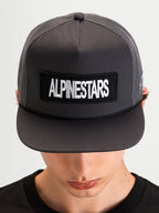 Alpinestars Cappello Ten, cappellino trucker sportswear a visiera piatta indossato da un modello, grigio scuro, con patch frontale con logo Alpinestars e corona in rete strutturata