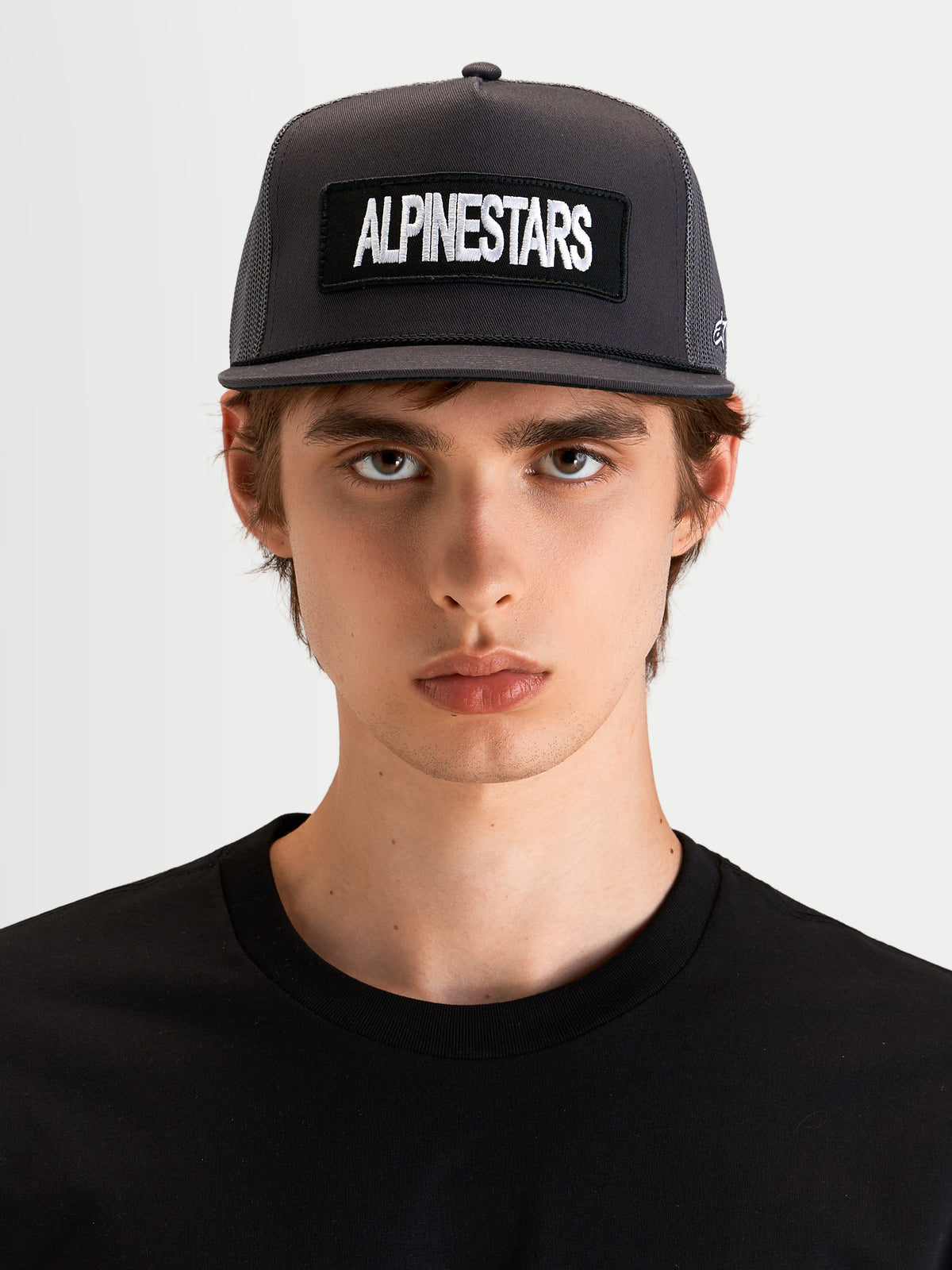 Alpinestars Cappello Ten, cappellino trucker sportswear a visiera piatta in grigio scuro, indossato da un modello, con corona strutturata e patch ricamata con logo Alpinestars sul davanti