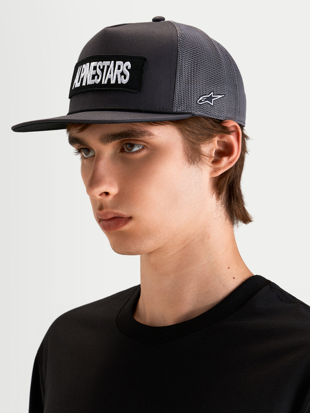 Alpinestars Cappello Ten, cappello trucker sportswear a visiera piatta in grigio scuro, indossato da un modello, con patch frontale con logo Alpinestars ricamato e ricamo laterale del logo Astars