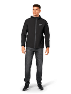 Giacca antipioggia Alpinestars Kascade, shell antipioggia da uomo, nero, indossata con jeans grigio scuro e sneakers nere, caratterizzata da design con cappuccio, cerniera frontale rivestita e logo Alpinestars bianco sul petto, streetwear casual ispirato al motorsport