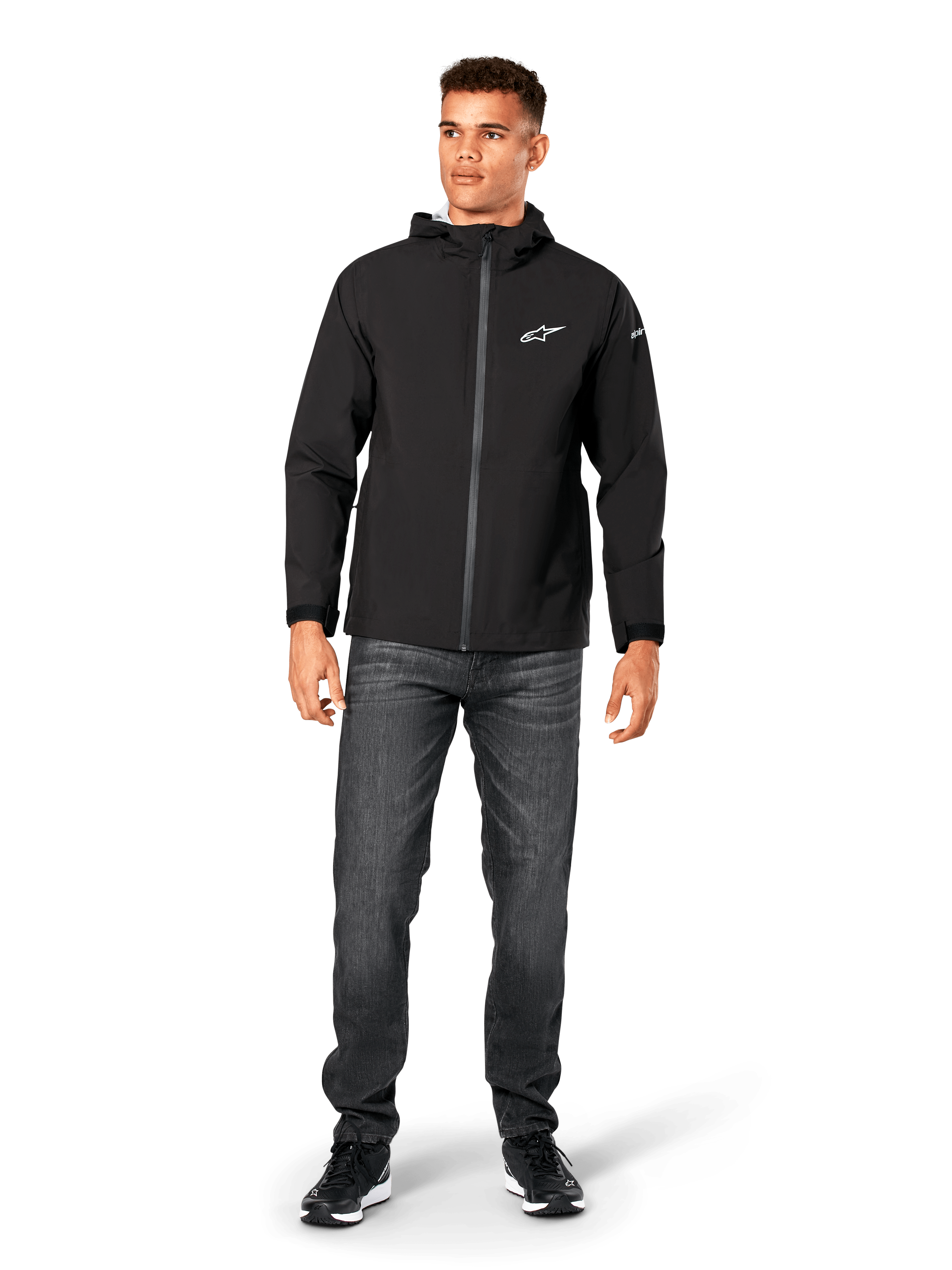 Giacca antipioggia Alpinestars Kascade, shell antipioggia da uomo, nero, indossata con jeans grigio scuro e sneakers nere, caratterizzata da design con cappuccio, cerniera frontale rivestita e logo Alpinestars bianco sul petto, streetwear casual ispirato al motorsport