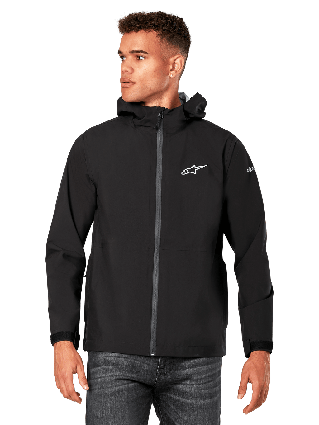 Modello che indossa la giacca antipioggia Alpinestars Kascade, nera, logo bianco sul petto e sulla manica, guscio antipioggia con cappuccio, zip frontale rivestita e polsini in velcro, abbinata a jeans scuri, streetwear casual ispirato al motorsport