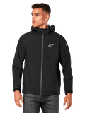 Modello che indossa la giacca antipioggia Alpinestars Kascade, nera, logo bianco sul petto e sulla manica, guscio antipioggia con cappuccio, zip frontale rivestita e polsini in velcro, abbinata a jeans scuri, streetwear casual ispirato al motorsport