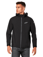 Modello che indossa la giacca antipioggia Alpinestars Kascade, nera, logo bianco sul petto e sulla manica, guscio antipioggia con cappuccio, zip frontale rivestita e polsini in velcro, abbinata a jeans scuri, streetwear casual ispirato al motorsport