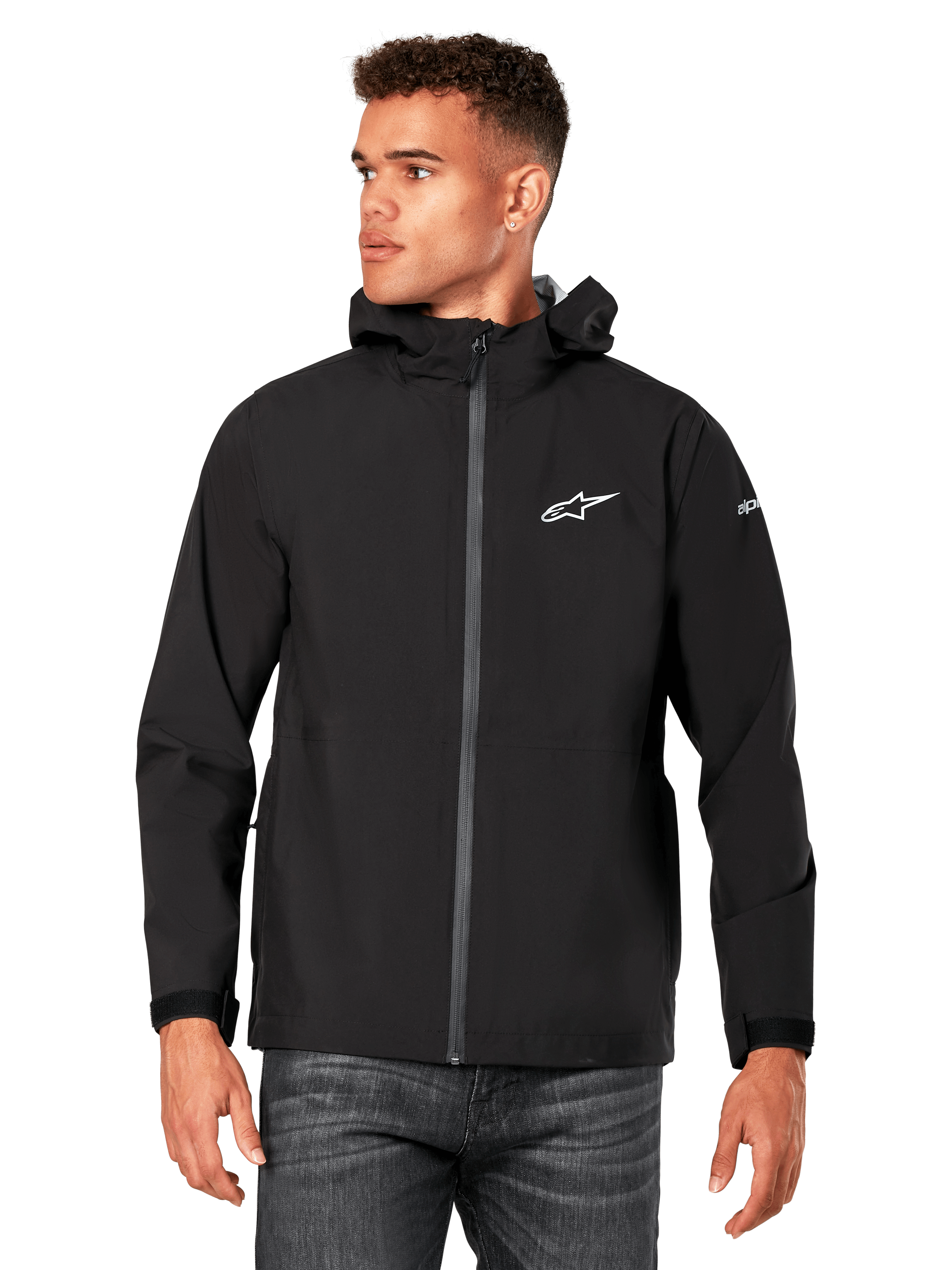 Modello che indossa la giacca antipioggia Alpinestars Kascade, nera, logo bianco sul petto e sulla manica, guscio antipioggia con cappuccio, zip frontale rivestita e polsini in velcro, abbinata a jeans scuri, streetwear casual ispirato al motorsport