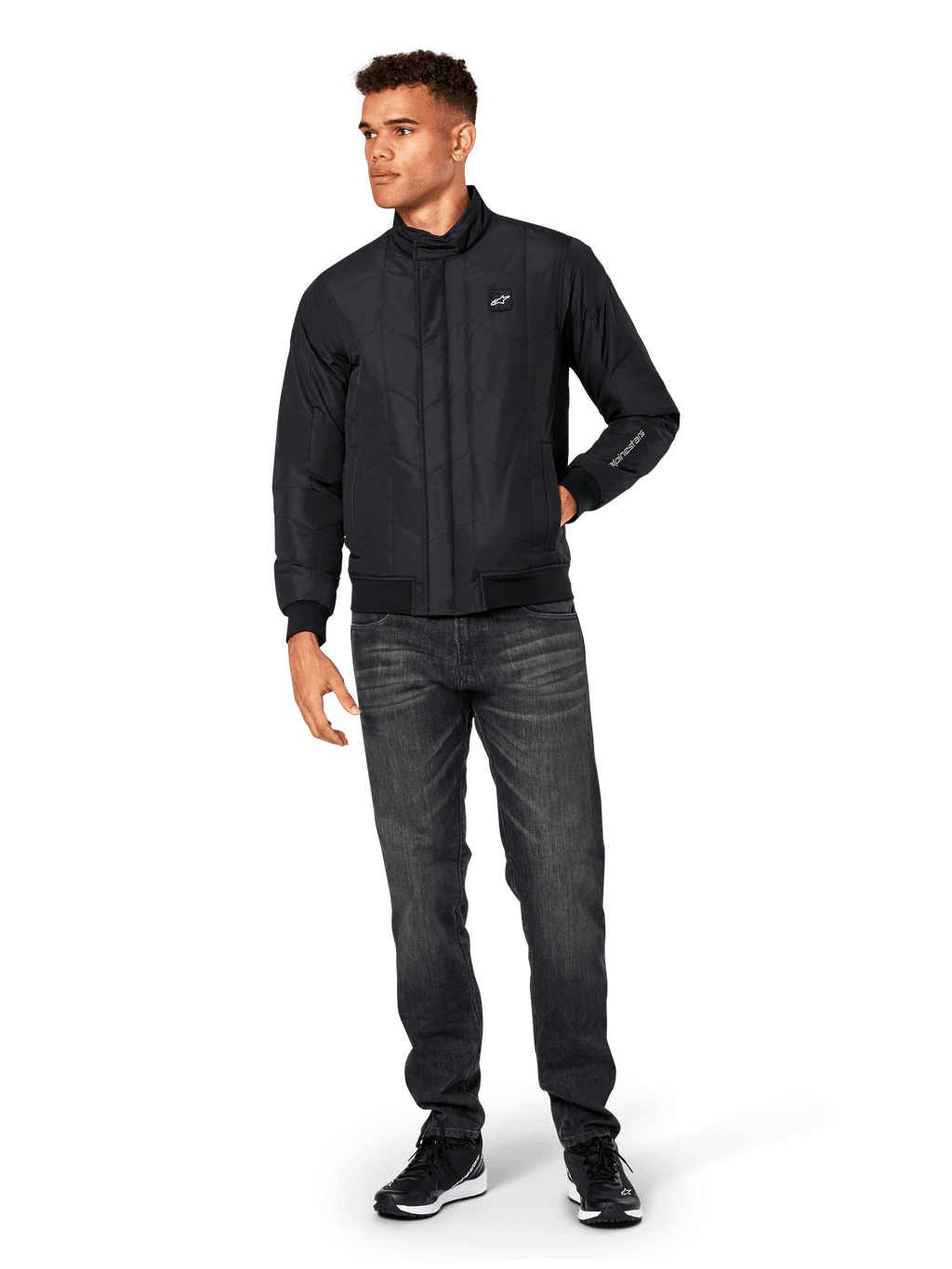 Giacca Alpinestars Precedent, giacca sportswear trapuntata, nera, indossata con jeans grigio scuro e sneakers nere, con motivo trapuntato angolato, colletto basso, polsini e orlo a coste, patch logo bianca sul petto e ricamo sulla manica.