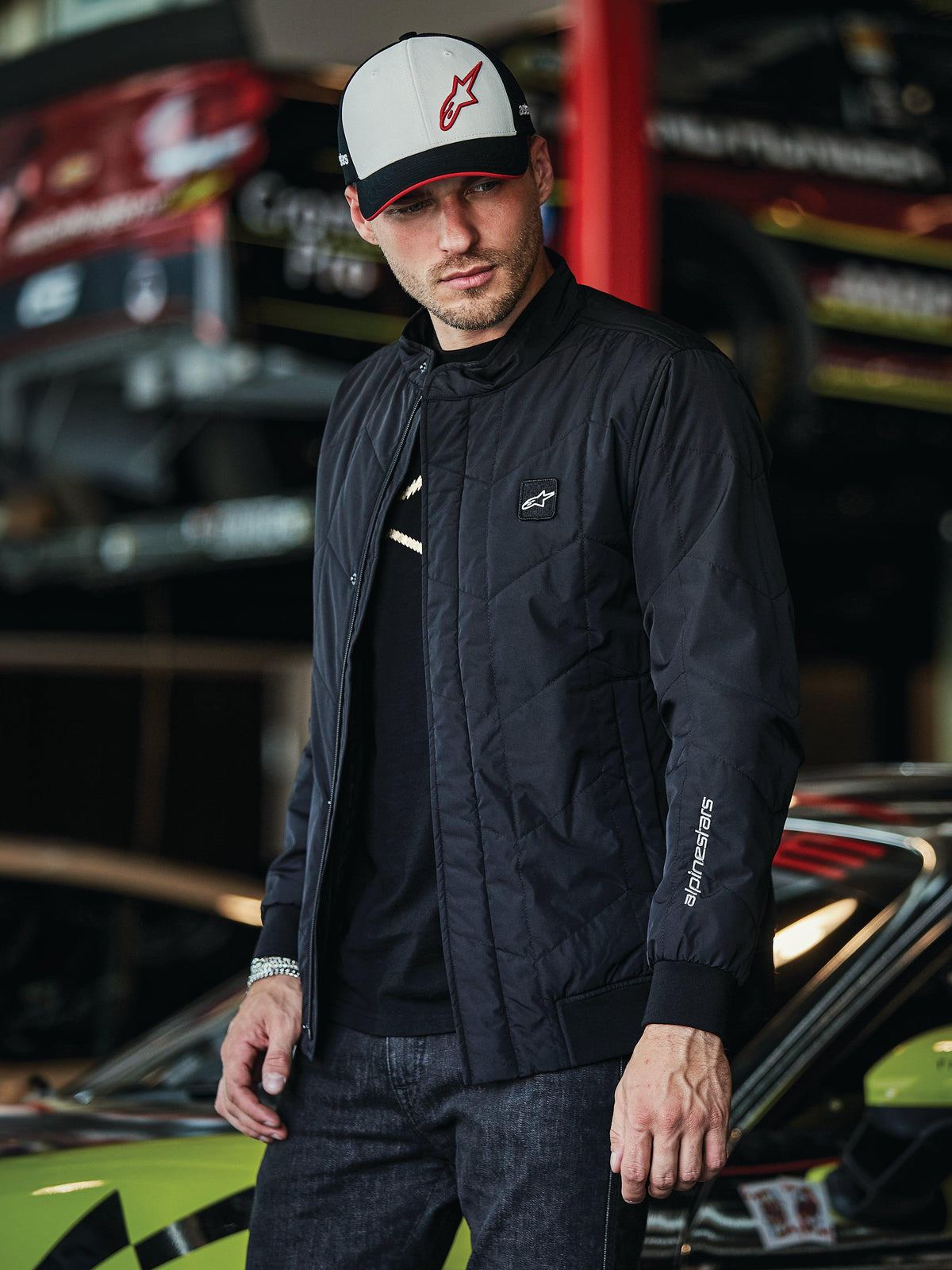 Alpinestars Precedent Jacket, giacca trapuntata nera con stile Formula 1, caratterizzata da un iconico motivo trapuntato angolato, patch con logo sul petto e ricamo bianco sulla manica, indossata con un cappellino Alpinestars coordinato in un garage di sport motoristici.