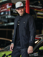 Alpinestars Precedent Jacket, giacca trapuntata nera con stile Formula 1, caratterizzata da un iconico motivo trapuntato angolato, patch con logo sul petto e ricamo bianco sulla manica, indossata con un cappellino Alpinestars coordinato in un garage di sport motoristici.